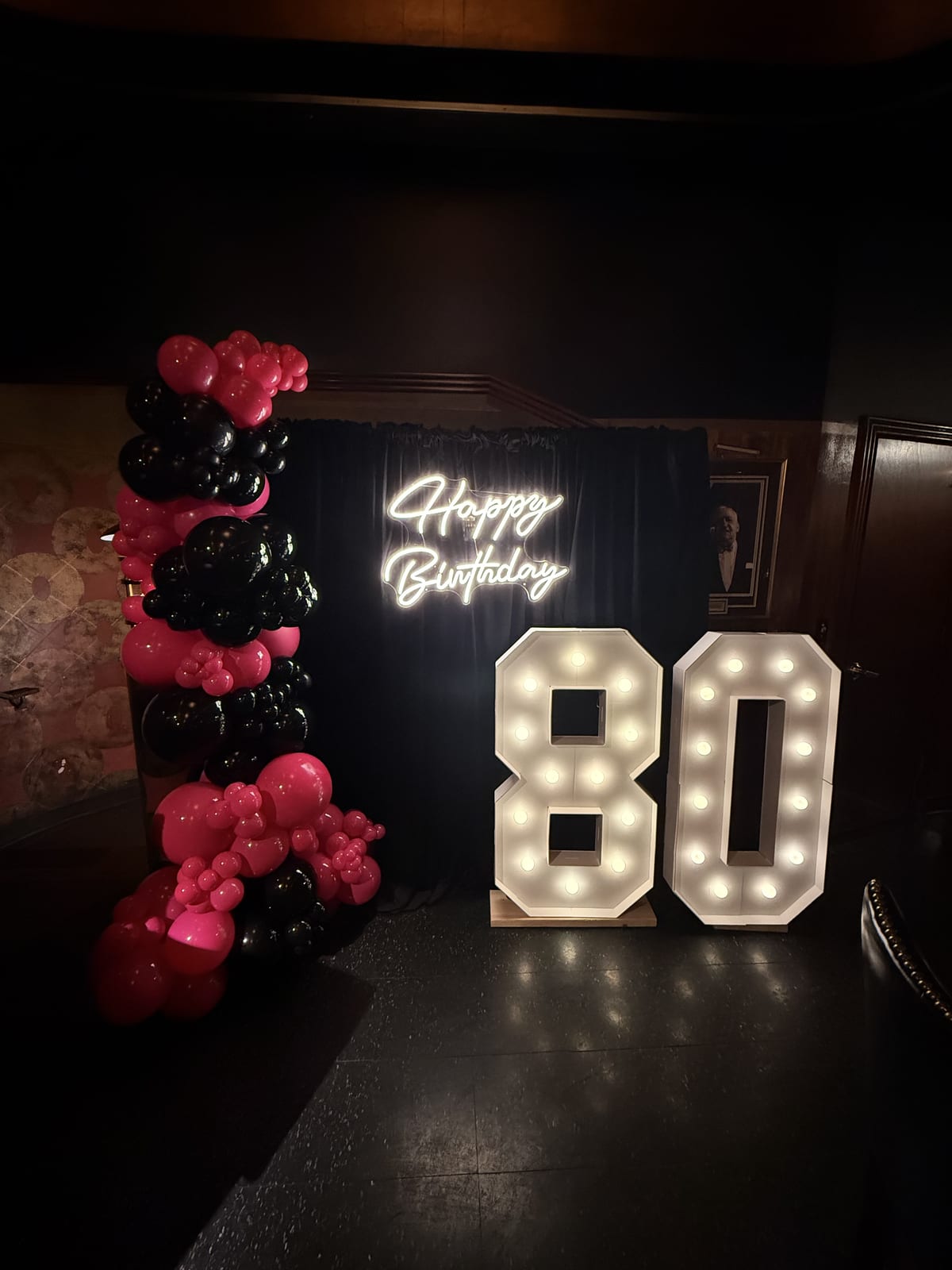 Marquee numbers 8 and 0 lit up at a birthday party with a balloon arch and a neon Happy Birthday sign